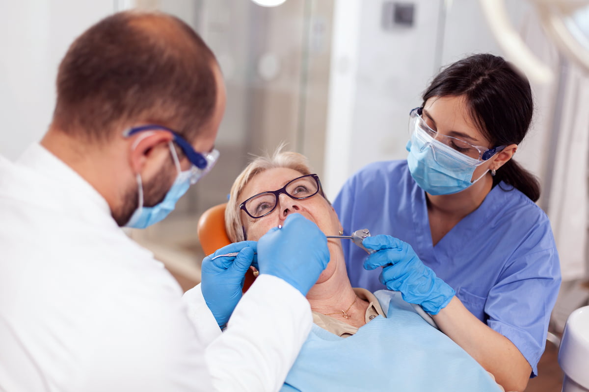Elderly woman receiving a dental examination from two dentists wearing masks and gloves.