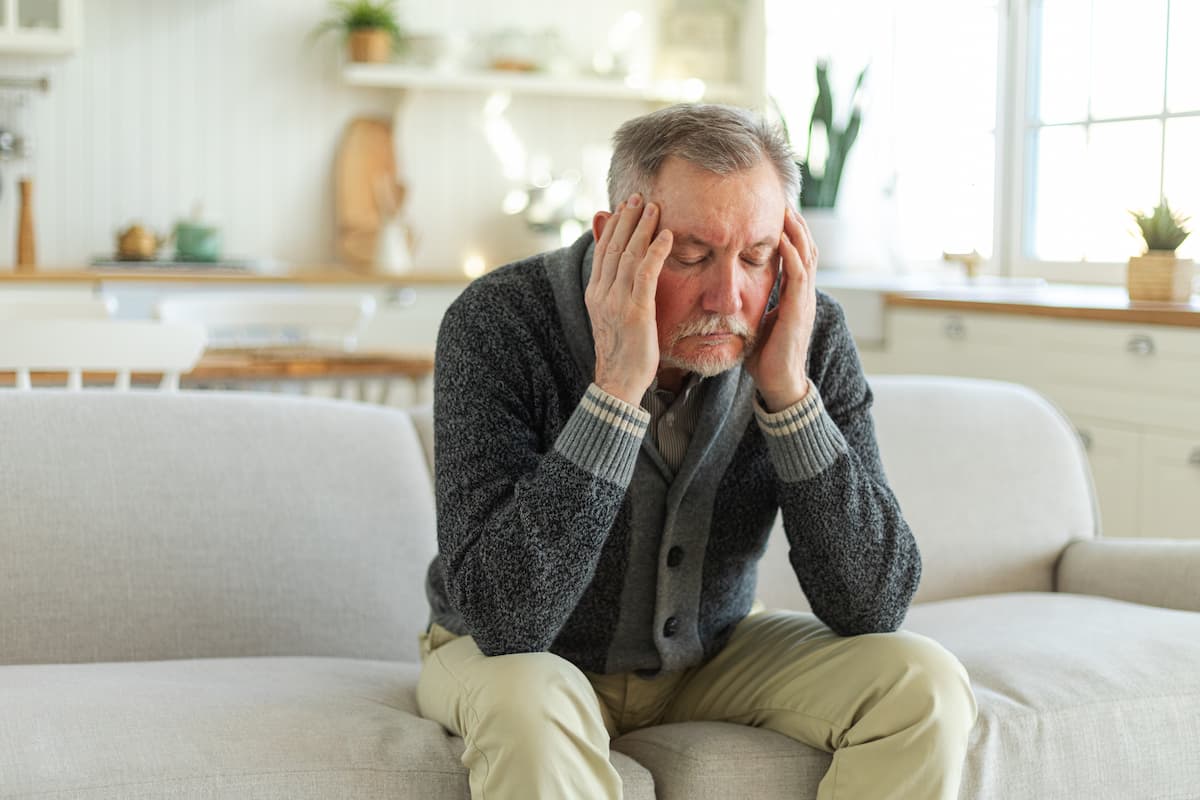 Elderly man sitting on a couch holding his head in stress after losing his dentures.
